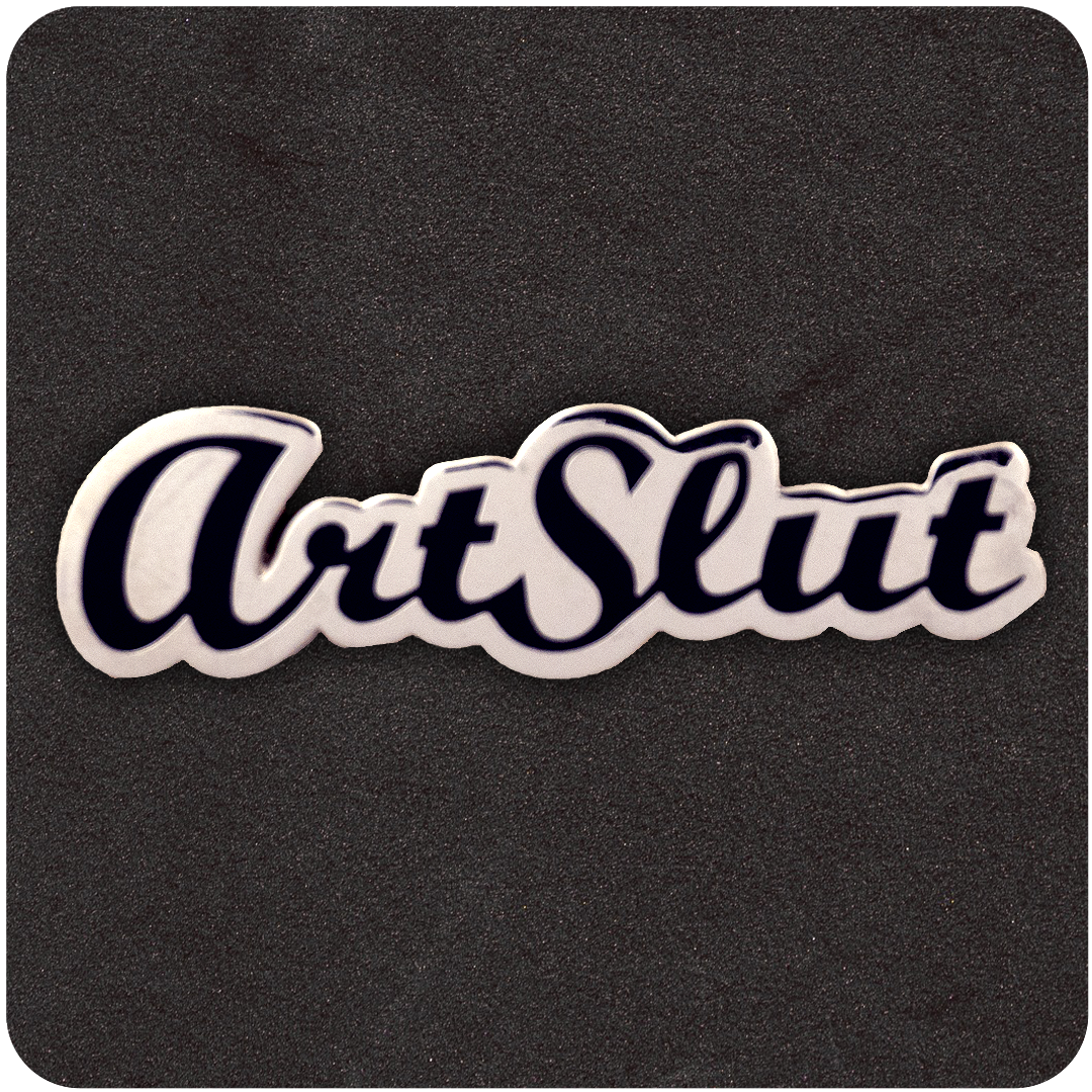 White and black enamel pin reading "ArtSlut" in bold, cursive script, displayed on a dark foam background.