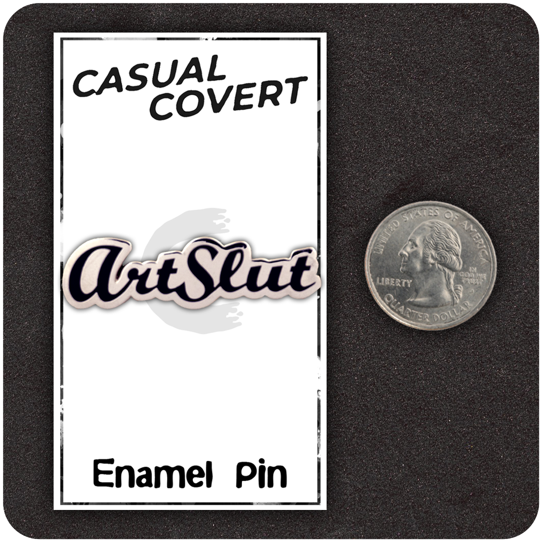 ArtSlut enamel pin on Casual Covert backing card shown next to a U.S. quarter for scale, featuring bold black script on a white pin, all placed on a black foam display.