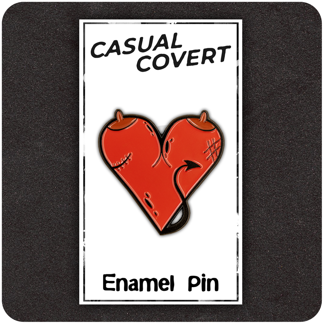 Red devil heart enamel pin with horns, stitches, and a tail, displayed on a branded “Casual Covert” backing card labeled “Enamel Pin,” set against a dark textured background.
