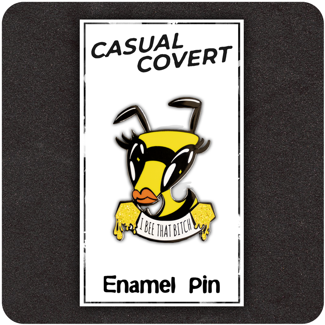 Funny enamel pin featuring a diva bee with bold lashes and orange lips, holding a dripping honey banner that says “I Bee That Bitch.” Displayed on a branded Casual Covert backing card against a dark foam background.