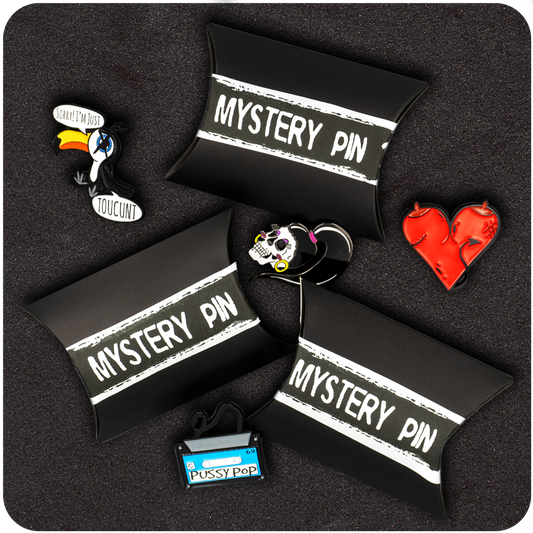 Four black pillow boxes labeled “Mystery Pin” arranged on a black textured background, surrounded by colorful enamel pins including a boxing glove heart, a sugar skull girl, a Pussy Pop cassette tape, and a toucan with a cheeky phrase.