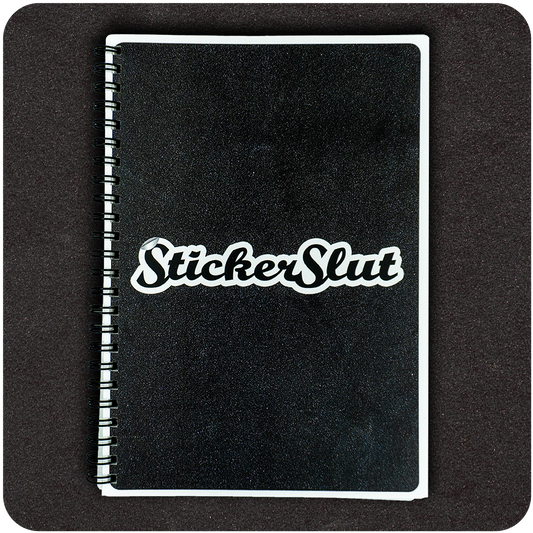 A black spiral-bound notebook with a glossy finish lies flat against a dark background. Centered on the front cover is a bold white and black vinyl sticker that reads “StickerSlut” in a playful, retro font. The minimalist, high-contrast design highlights the sticker and gives the book a cheeky, rebellious vibe.