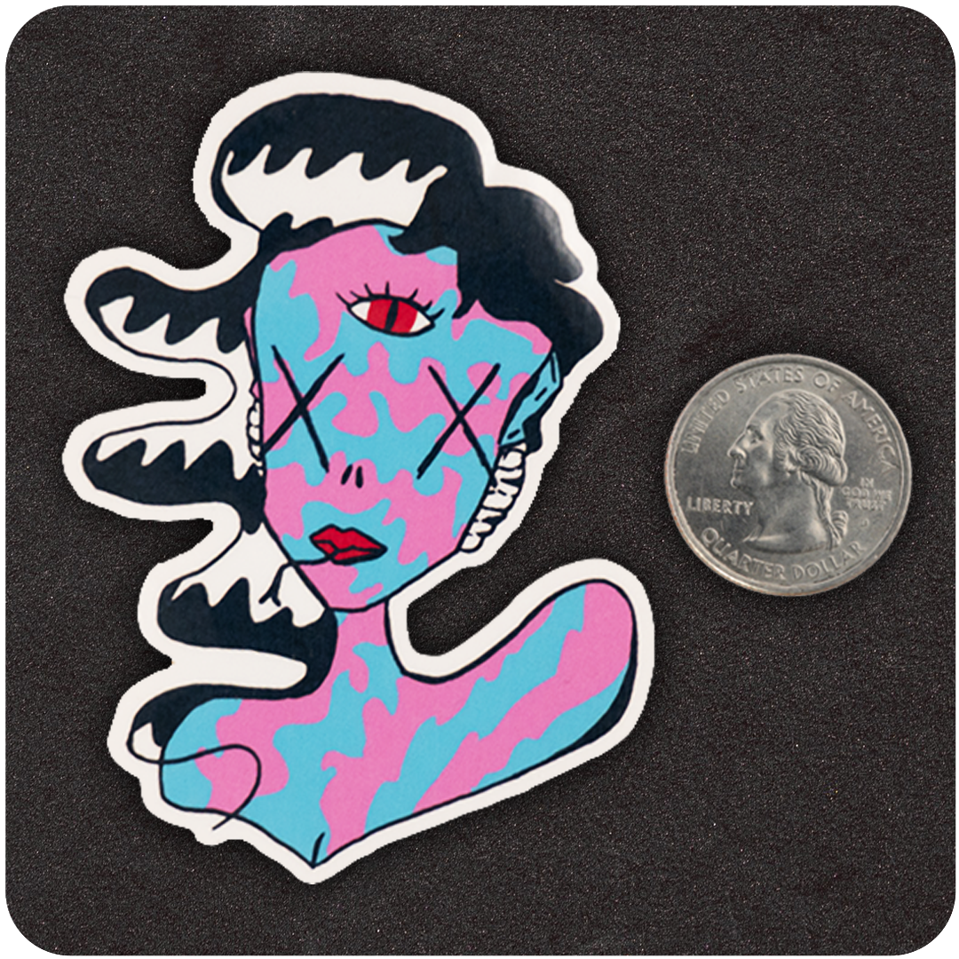 A colorful die-cut vinyl sticker of a surreal character featuring swirling black-and-white hair, a camouflage-like pink and blue face and body, X’s over the eyes, and a striking third eye in red on the forehead. The figure also has red lips and an abstract expression. Positioned next to a U.S. quarter for scale, the sticker rests on a textured black background, highlighting its bold, vibrant design and medium size.