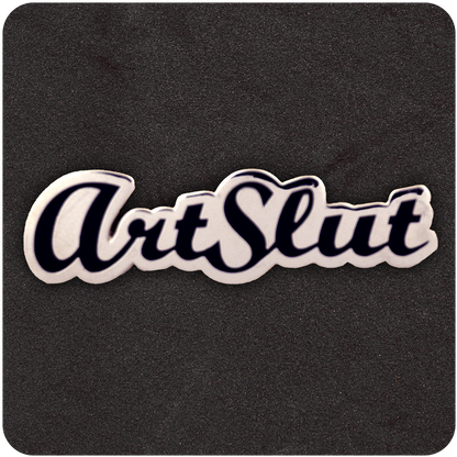 White and black enamel pin reading "ArtSlut" in bold, cursive script, displayed on a dark foam background.