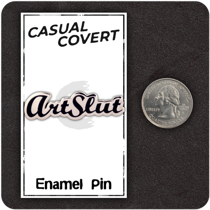 ArtSlut enamel pin on Casual Covert backing card shown next to a U.S. quarter for scale, featuring bold black script on a white pin, all placed on a black foam display.