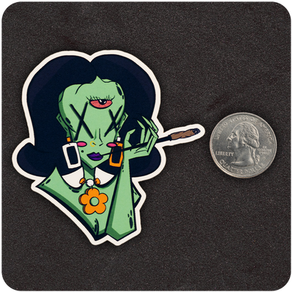 A vinyl sticker of a green alien woman with three eyes—two marked with black Xs and a third in the center of her forehead. She has large black hair, geometric earrings (one white, one orange), and wears a white collared top with an orange flower necklace. Her lips are purple, and she’s holding a lit joint in one hand. A U.S. quarter is placed beside the sticker for scale on a textured black background.