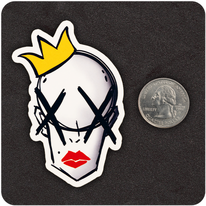 A die-cut vinyl sticker featuring a stylized illustration of a bald head with a yellow crown, bold red lips, and large black Xs over the eyes. The sticker is placed on a dark surface next to a U.S. quarter, showcasing its compact size. The clean, edgy design contrasts sharply with the black background, emphasizing its vibrant colors and graphic details.