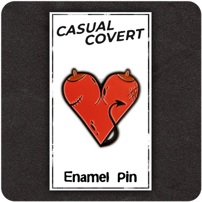 Red devil heart enamel pin with horns, stitches, and a tail, displayed on a branded “Casual Covert” backing card labeled “Enamel Pin,” set against a dark textured background.