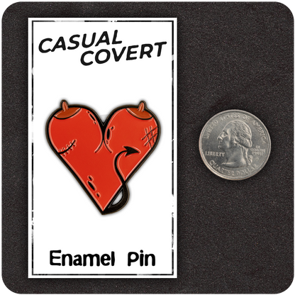 Red devil heart enamel pin with horns and a tail, shown on a Casual Covert branded backing card next to a U.S. quarter for size reference, set on a dark textured background.