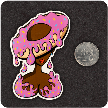 A vinyl sticker featuring a stylized woman with brown skin whose head and chest are partially covered in dripping pink frosting with colorful sprinkles. Her head resembles a doughnut with a hole in the center, and the frosting drips over her closed eyes and onto her chest, which is also covered in pink icing. A U.S. quarter is placed to the right of the sticker for size reference. The background is a dark textured surface.