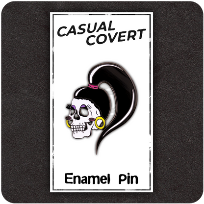 Drag Me To Hell enamel pin on branded Casual Covert backing card, featuring a femme skull with exaggerated eyelashes, purple eyeshadow, yellow hoop earrings, and a long black ponytail.