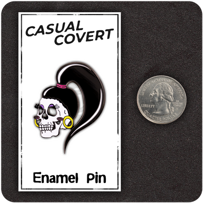 Drag Me To Hell enamel pin displayed on a Casual Covert backing card next to a U.S. quarter for scale, featuring a femme skull with bold makeup, yellow hoop earrings, and a high black ponytail.