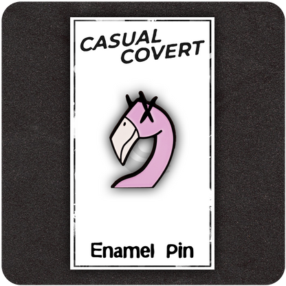 Pink flamingo enamel pin with cartoon X eyes, mounted on a white backing card labeled "Casual Covert" and "Enamel Pin," set against a dark background.