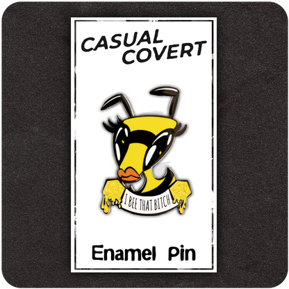 Funny enamel pin featuring a diva bee with bold lashes and orange lips, holding a dripping honey banner that says “I Bee That Bitch.” Displayed on a branded Casual Covert backing card against a dark foam background.