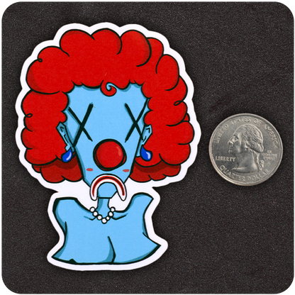 A vinyl sticker of a blue-skinned cartoon clown bust with a bright red afro, oversized red nose, Xs for eyes, blue earrings, a pearl necklace, and a deep frown. Positioned next to a U.S. quarter for size reference, the sticker is shown on a textured black background.