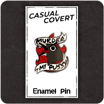 Enamel pin of a cartoon bird with red glitter flames and a bold banner that reads “Murder My Pussy,” displayed on a white Casual Covert backing card with a black foam background.