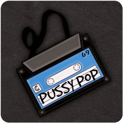 Pussy Pop Mixtape enamel pin shaped like a retro cassette tape with a blue label, black trim, and black tape unraveling, displayed on a black foam background.