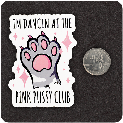 A die-cut vinyl sticker placed next to a U.S. quarter for scale. The sticker shows a cartoon-style raised paw with pink pads and claws, surrounded by sparkling pink stars. The top text says “I’M DANCIN AT THE” and the bottom reads “PINK PUSSY CLUB.” Set against a black background.