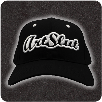 Front view of a black snapback hat with bold white embroidered “Art Slut” logo, centered on a structured crown and displayed against a dark textured backdrop with soft halo lighting.