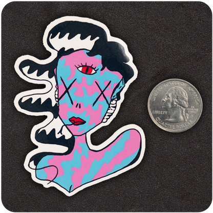 A colorful die-cut vinyl sticker of a surreal character featuring swirling black-and-white hair, a camouflage-like pink and blue face and body, X’s over the eyes, and a striking third eye in red on the forehead. The figure also has red lips and an abstract expression. Positioned next to a U.S. quarter for scale, the sticker rests on a textured black background, highlighting its bold, vibrant design and medium size.